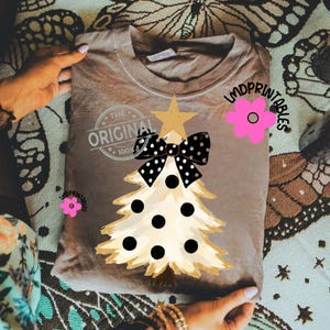 May include: A tan-colored t-shirt featuring a Christmas tree design. The tree is cream-colored with black polka dots and a black and white polka dot bow. The text "THE ORIGINAL 100%" is printed on the shirt.
