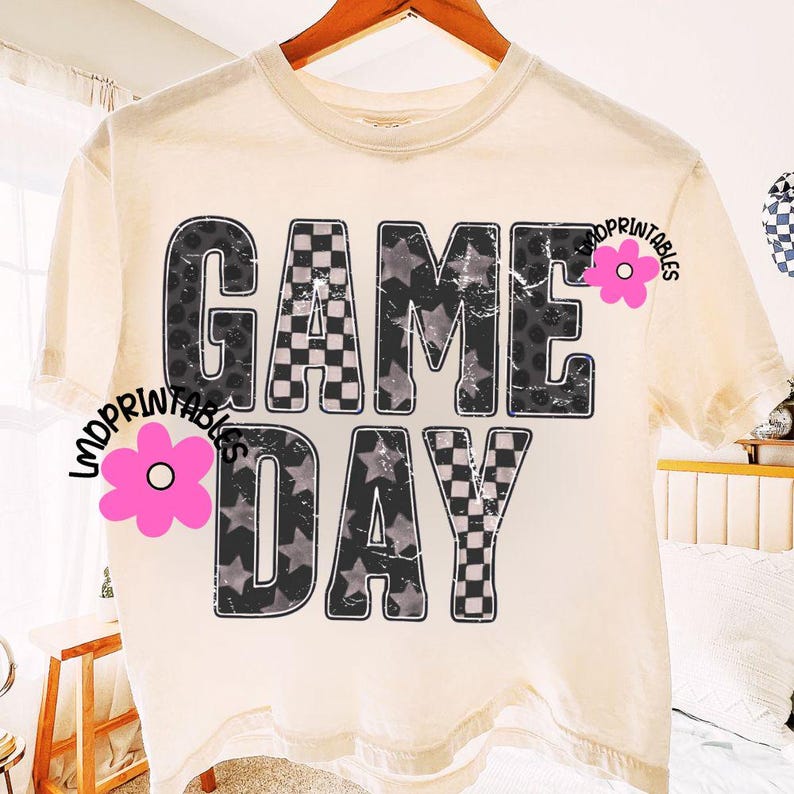 May include: Cream-colored t-shirt with the words "GAME DAY" in large, distressed black letters. The letters are filled with patterns like stars, checkered squares, and leopard print. A casual, comfortable tee.
