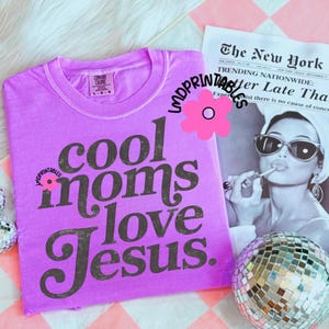 Puede incluir: Una camiseta de color lila con el texto "cool moms love Jesus" en una fuente oscura y vintage. La camiseta se muestra con una imagen en blanco y negro de una mujer y una bola de discoteca.