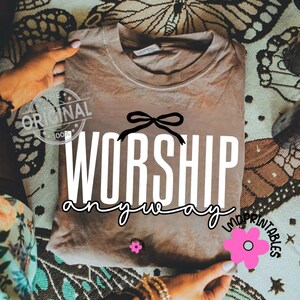 May include: A folded brown t-shirt with the words "WORSHIP anyway" in white script and a black bow graphic. The shirt has a vintage look and is held by hands. A pink flower graphic and the text "LMPRINTABLES" are also visible.