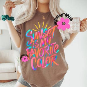 May include: A brown t-shirt with the text "Sunset is my favorite color" in colorful, stylized lettering. The design includes a yellow sunburst and the words are in shades of blue, pink, orange, and green.