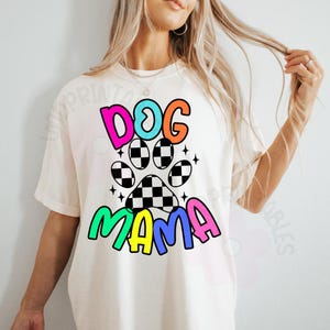 May include: Cream-colored t-shirt with the words "DOG MAMA" in colorful, bubble letters. The design includes a paw print with a checkered pattern and star accents, creating a playful and vibrant aesthetic. A casual, comfortable style.