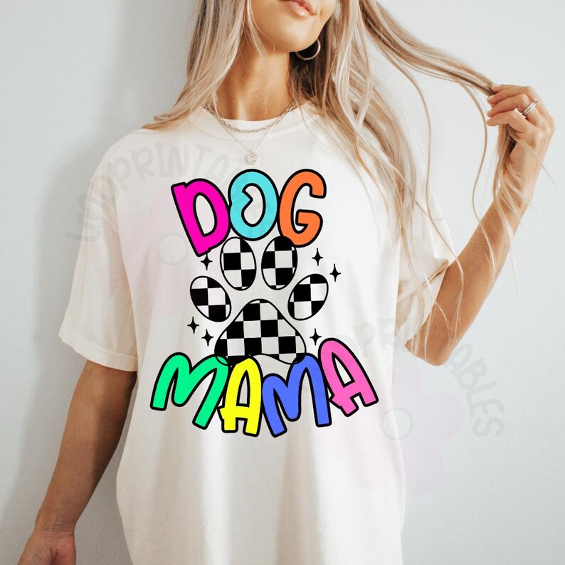 May include: Cream-colored t-shirt with the words "DOG MAMA" in colorful, bubble letters. The design includes a paw print with a checkered pattern and star accents, creating a playful and vibrant aesthetic. A casual, comfortable style.