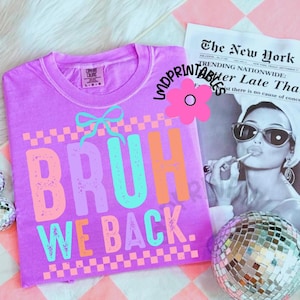 Puede incluir: Una camiseta morada con el texto "BRUH WE BACK" en letras coloridas. La camiseta tiene un lazo gr&aacute;fico encima de la palabra "BRUH". Un recorte de peri&oacute;dico y una bola de discoteca est&aacute;n en el fondo.