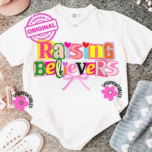 May include: White t-shirt with the text "Raising Believers" in colorful, block letter style. The design includes a pink bow and a pink flower graphic. A pink "Original" stamp is visible.