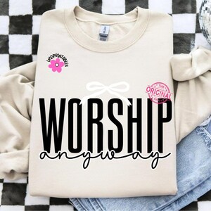 May include: A cream-coloured sweatshirt featuring the words "WORSHIP anyway" in black script. A pink flower graphic with the text "indprintables" is on the left, and a pink stamp that says "THE ORIGINAL 100%" is on the right. A white bow is above the word "WORSHIP".