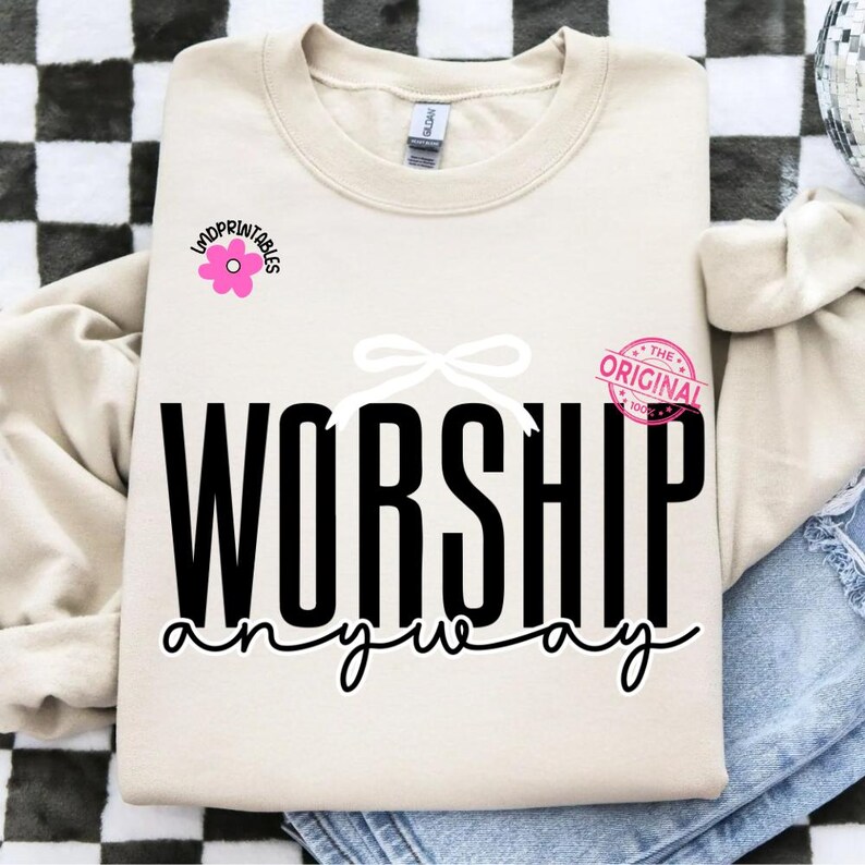 May include: A cream-coloured sweatshirt featuring the words "WORSHIP anyway" in black script. A pink flower graphic with the text "indprintables" is on the left, and a pink stamp that says "THE ORIGINAL 100%" is on the right. A white bow is above the word "WORSHIP".