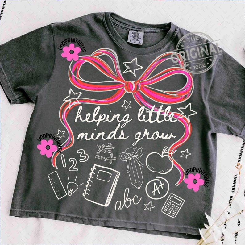 May include: Dark gray t-shirt with a large pink and red bow design and the words "helping little minds grow." The shirt features school-themed graphics like an apple, pencil, and notebook. Pink flower accents are on the shirt.