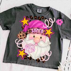Op de afbeelding: Donkergrijs t-shirt met een kerstman-afbeelding. De kerstman draagt een roze hoed met het woord "SANTA" en een luipaardprint. Gele sterren, roze krullen en de tekst "THE ORIGINAL 100%" maken het ontwerp af.