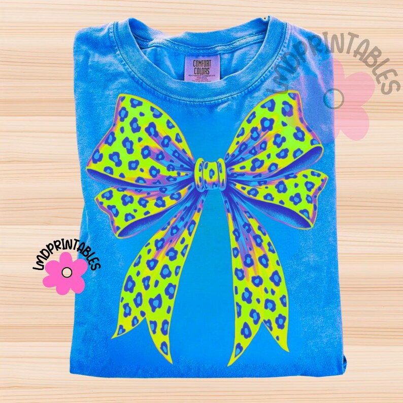 May include: A blue Comfort Colors t-shirt featuring a large bow design. The bow has a neon green and blue leopard print pattern with purple accents. The shirt is folded and displayed on a light wood surface. The text "LMDPrintables" is visible.
