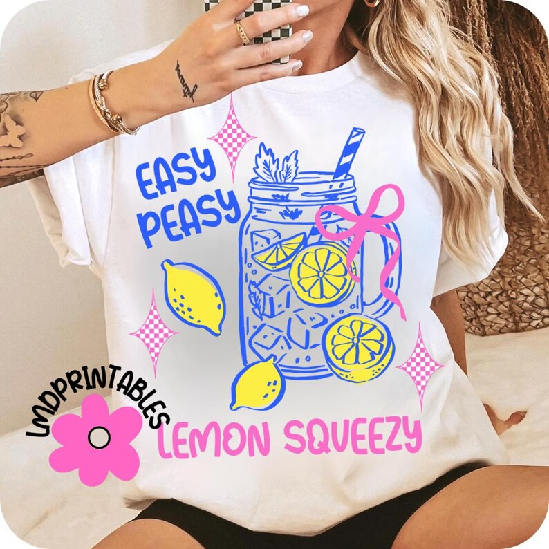 May include: White t-shirt with a colourful graphic design featuring a mason jar filled with lemons and ice cubes, the words "Easy Peasy Lemon Squeezy", and pink starburst accents. The shirt has short sleeves.