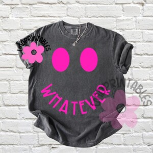May include: A charcoal gray t-shirt with a smiley face design. The smiley face features two large pink ovals for eyes and the word "WHATEVER" in pink as the mouth. A silver necklace is visible at the neckline.