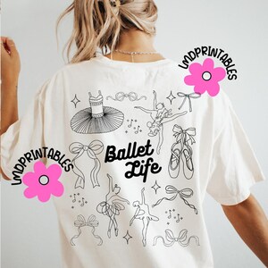 May include: Cream-colored t-shirt featuring a black line art design of ballet-themed elements. The design includes a tutu, ballet shoes, bows, dancers, and musical notes. The words "Ballet Life" are also printed on the shirt.
