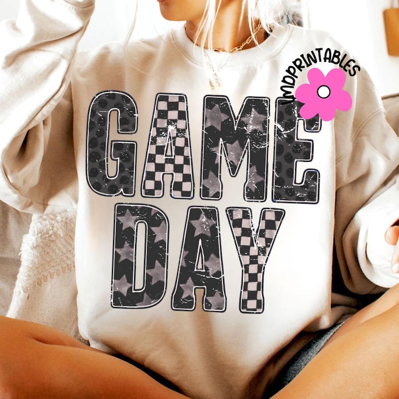 May include: Cream-colored sweatshirt with the words "GAME DAY" in large, distressed black lettering. The letters are filled with checkered, star, and polka dot patterns. A pink flower logo is visible in the upper right corner.