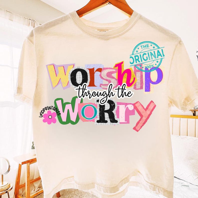 May include: Cream-colored t-shirt with the words "Worship through the Worry" in a colorful, layered design. The text is in various fonts and colors, including yellow, pink, and green. A circular stamp reads "The Original 100%."