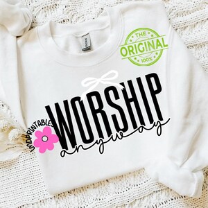 May include: White sweatshirt featuring the text "WORSHIP anyway" in black. A white bow and a pink flower graphic are above and to the left of the text. The words "THE ORIGINAL 100%" are printed in a green circle.