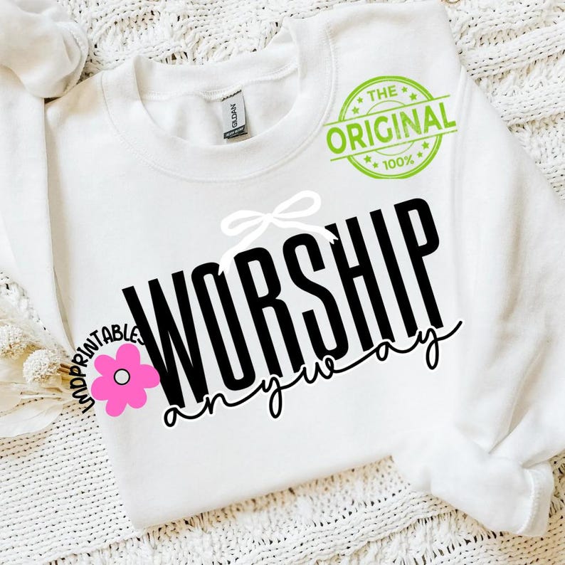 May include: White sweatshirt featuring the text "WORSHIP anyway" in black. A white bow and a pink flower graphic are above and to the left of the text. The words "THE ORIGINAL 100%" are printed in a green circle.