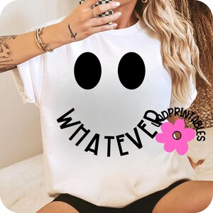 May include: White t-shirt featuring a large smiley face design with black eyes and the word "WHATEVER" in black text forming the mouth. A pink flower graphic is on the right side of the mouth. The shirt is a casual, everyday wear item.