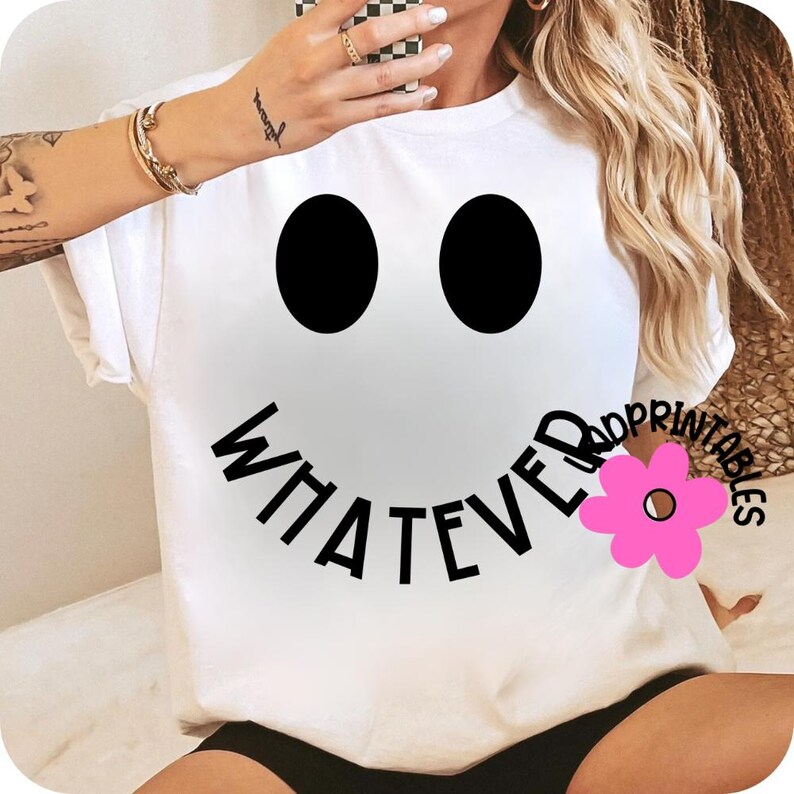 May include: White t-shirt featuring a large smiley face design with black eyes and the word "WHATEVER" in black text forming the mouth. A pink flower graphic is on the right side of the mouth. The shirt is a casual, everyday wear item.