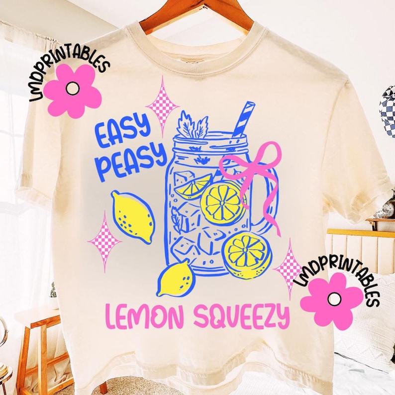 May include: A cream-coloured t-shirt featuring a blue and yellow lemonade design. The graphic shows a mason jar with ice, lemons, and a straw, along with the words "Easy Peasy Lemon Squeezy".