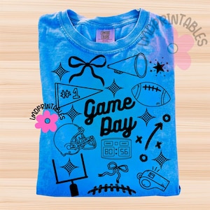 May include: A light blue t-shirt with a graphic design featuring football-themed elements. The design includes a football, helmet, megaphone, and the words "Game Day". The shirt also has a score display and a whistle.