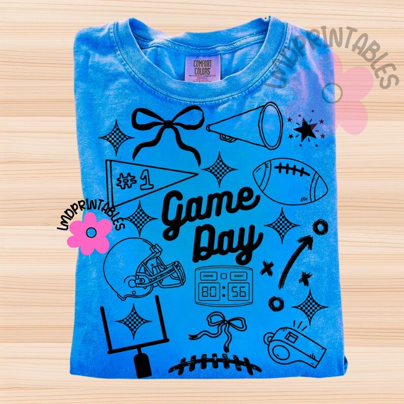 May include: A light blue t-shirt with a graphic design featuring football-themed elements. The design includes a football, helmet, megaphone, and the words "Game Day". The shirt also has a score display and a whistle.