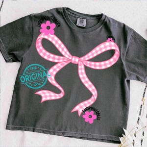 May include: Dark gray t-shirt featuring a large pink gingham bow graphic with pink flowers and the text "LMDPRINTABLES". The shirt also has a teal circular graphic that says "THE ORIGINAL 100%."