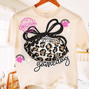May include: Cream-colored t-shirt featuring a football with a leopard print design and a black bow. The text "gameday" is written in a cursive font. The shirt also has the text "THE ORIGINAL 100%" and the word "MDPRINTABLES" in pink.
