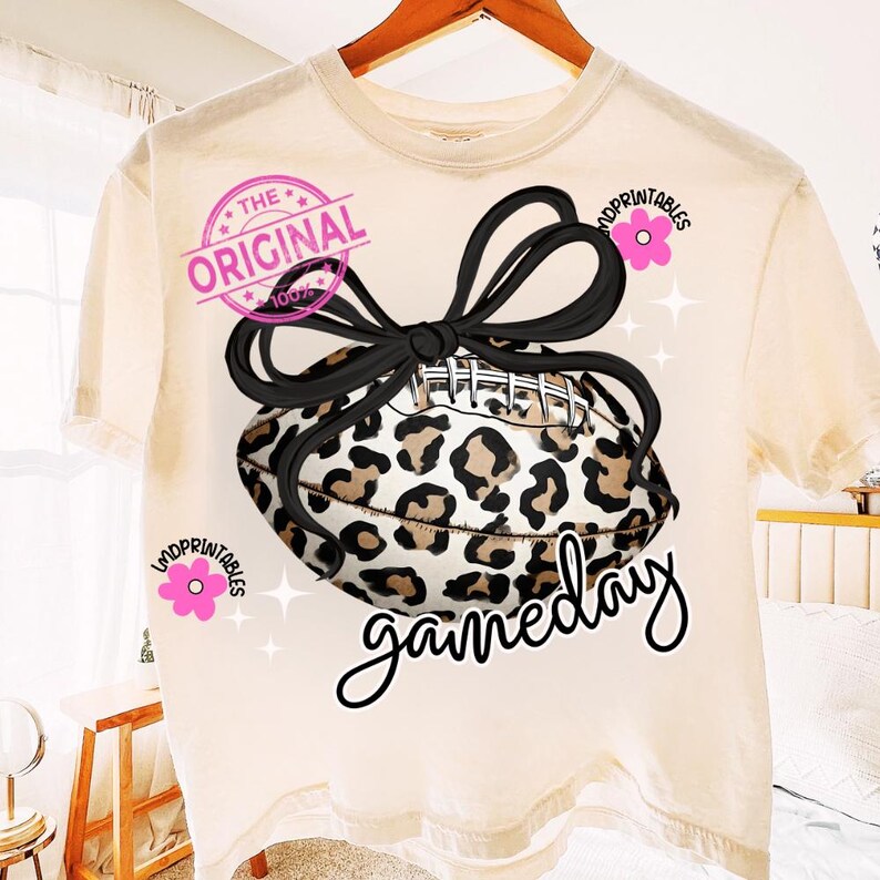 May include: Cream-colored t-shirt featuring a football with a leopard print design and a black bow. The text "gameday" is written in a cursive font. The shirt also has the text "THE ORIGINAL 100%" and the word "MDPRINTABLES" in pink.