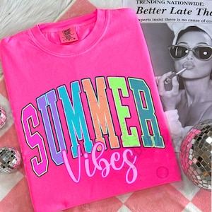 Puede incluir: Camiseta rosa brillante Comfort Colors con las palabras "SUMMER Vibes" en una fuente retro colorida. Las letras est&aacute;n delineadas en varios colores, incluyendo morado, azul, naranja y verde. La camiseta se muestra sobre una superficie a cuadros rosa y blanco.