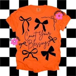 K&ouml;nnte beinhalten: Leuchtend orangefarbenes T-Shirt mit schwarzen Schleifenmotiven und dem Schriftzug "Count Your Blessings" in Schreibschrift. Das Shirt hat auch eine rosa Blumengrafik und den Text "LMDPRINTABLES".