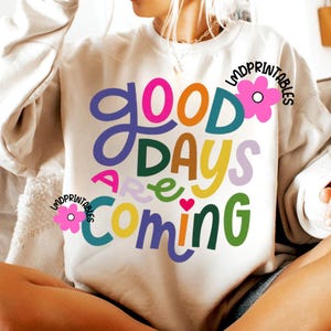 Może przedstawiać: Kremowa bluza z napisem "Good Days Are Coming" w kolorowych, zabawnych literach. Projekt zawiera r&oacute;żowy, pomarańczowy, zielony, fioletowy i ż&oacute;łty tekst z małym czerwonym sercem. Bluza ma okrągły dekolt i długie rękawy.