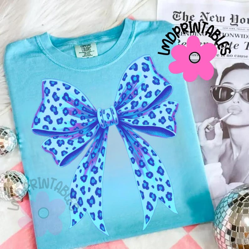 May include: A light blue t-shirt featuring a large bow design with a leopard print pattern in shades of blue and purple. The shirt has a crew neck and short sleeves. The text "LMD PRINTABLES" is printed on the shirt.