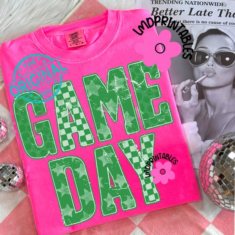 May include: A bright pink t-shirt with the words "GAME DAY" in large, distressed green letters. The letters have a checkered pattern and star accents. The shirt also features the text "THE ORIGINAL" in a teal circle.