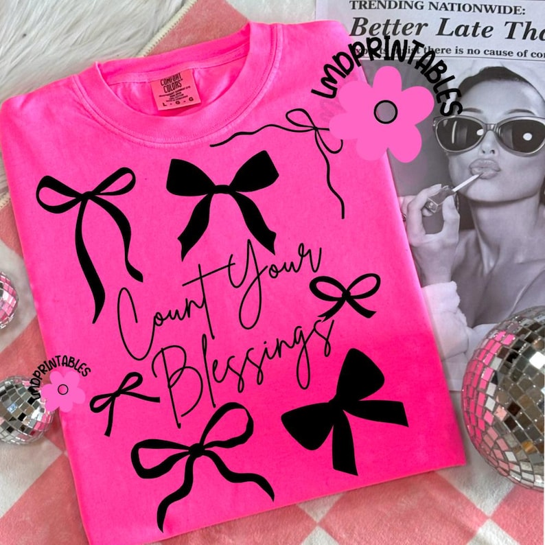 K&ouml;nnte beinhalten: Ein leuchtend pinkfarbenes Sweatshirt mit schwarzen Schleifenmotiven und dem Schriftzug "Count Your Blessings" in Schreibschrift. Das Kleidungsst&uuml;ck hat einen Rundhalsausschnitt und lange &Auml;rmel. Das Etikett "Comfort Colors" ist sichtbar.
