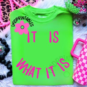 May include: A neon green t-shirt with the phrase "IT IS WHAT IT IS" in bright pink lettering. A pink flower graphic is above the words "IT IS". The shirt is a Gildan brand.