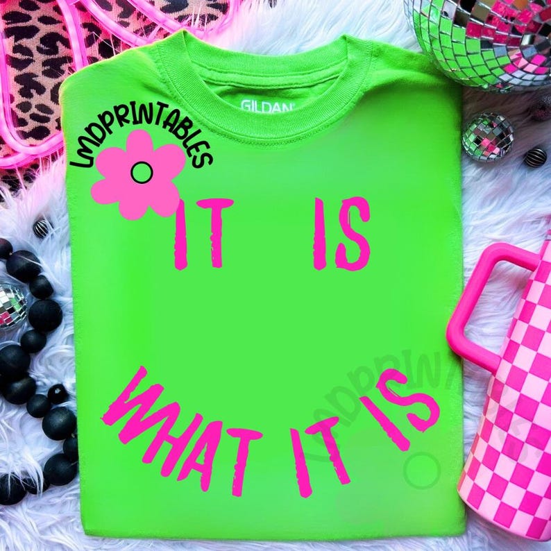 May include: A neon green t-shirt with the phrase "IT IS WHAT IT IS" in bright pink lettering. A pink flower graphic is above the words "IT IS". The shirt is a Gildan brand.