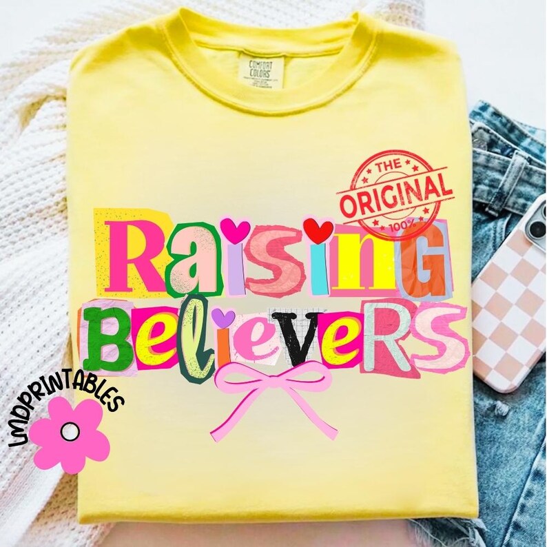 May include: A yellow t-shirt with the words "Raising Believers" in a colorful, cut-out style design. The shirt also features a pink bow, a flower graphic, and a red stamp that says "The Original".