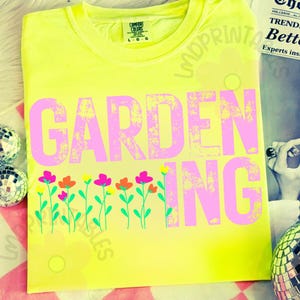 May include: A neon yellow t-shirt with the word "GARDENING" in large, distressed pink letters. Below the text is a row of colorful flowers with green stems. The shirt has a crew neck and short sleeves.