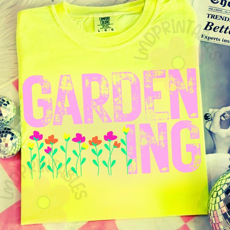 May include: A neon yellow t-shirt with the word "GARDENING" in large, distressed pink letters. Below the text is a row of colorful flowers with green stems. The shirt has a crew neck and short sleeves.