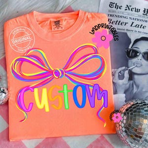 May include: Coral-colored t-shirt with a large, colorful bow design and the word "CUSTOM" in rainbow hues. The shirt has a round neckline and short sleeves. The text "ORIGINAL" is printed on the shirt. A newspaper and disco ball are in the background.