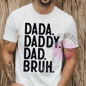 May include: White t-shirt with the words "DADA. DADDY. DAD. BRUH." printed in bold black letters. The shirt is a casual style, suitable for everyday wear. The design is simple and direct, making a statement.