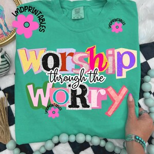 May include: A teal Comfort Colors t-shirt with the phrase "Worship Through the Worry" in a colorful, cut-out style. The word "Worship" is in yellow, pink, and purple. The word "Worry" is in pink and black. Pink flower graphics are on the shirt.
