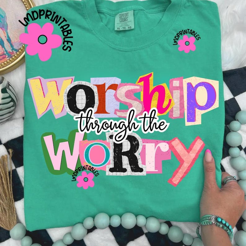 May include: A teal Comfort Colors t-shirt with the phrase "Worship Through the Worry" in a colorful, cut-out style. The word "Worship" is in yellow, pink, and purple. The word "Worry" is in pink and black. Pink flower graphics are on the shirt.