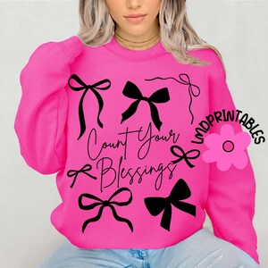 K&ouml;nnte beinhalten: Ein leuchtend pinkfarbenes Sweatshirt mit schwarzen Schleifenmotiven und dem Schriftzug "Count Your Blessings" in Schreibschrift. Das Design umfasst mehrere schwarze Schleifen in verschiedenen Stilen. Das Sweatshirt hat lange &Auml;rmel.