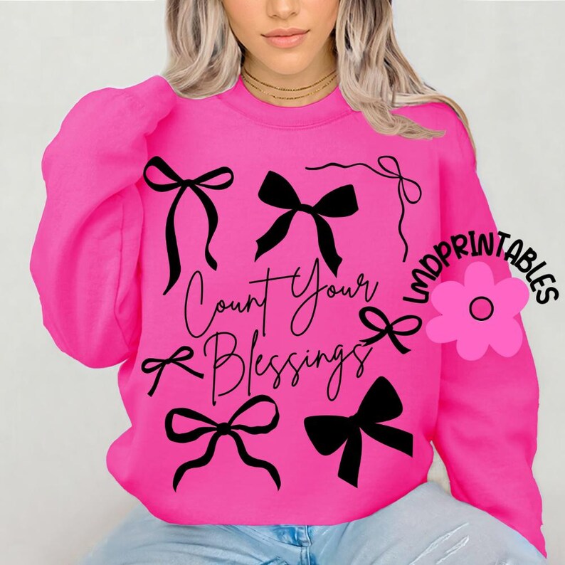K&ouml;nnte beinhalten: Ein leuchtend pinkfarbenes Sweatshirt mit schwarzen Schleifenmotiven und dem Schriftzug "Count Your Blessings" in Schreibschrift. Das Design umfasst mehrere schwarze Schleifen in verschiedenen Stilen. Das Sweatshirt hat lange &Auml;rmel.