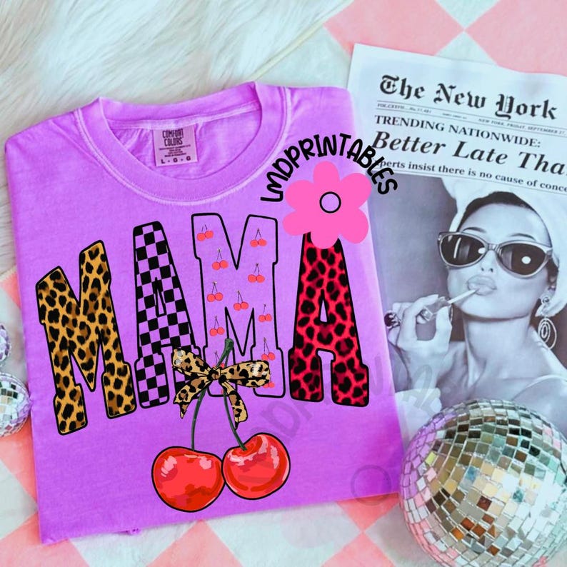 May include: A lilac-colored t-shirt with the word "MAMA" in a mix of leopard print, checkered, and cherry designs. Two red cherries with a bow are below the word. The shirt is from Comfort Colors.