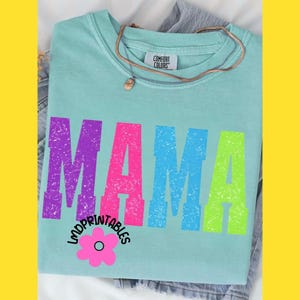 Pu&ograve; includere: Una maglietta Comfort Colors azzurra con la scritta "MAMA" a grandi lettere colorate e invecchiate. La maglietta presenta anche il testo "LMDPRINTABLES" e una grafica floreale rosa. Una collana d'oro &egrave; appoggiata sulla maglietta.