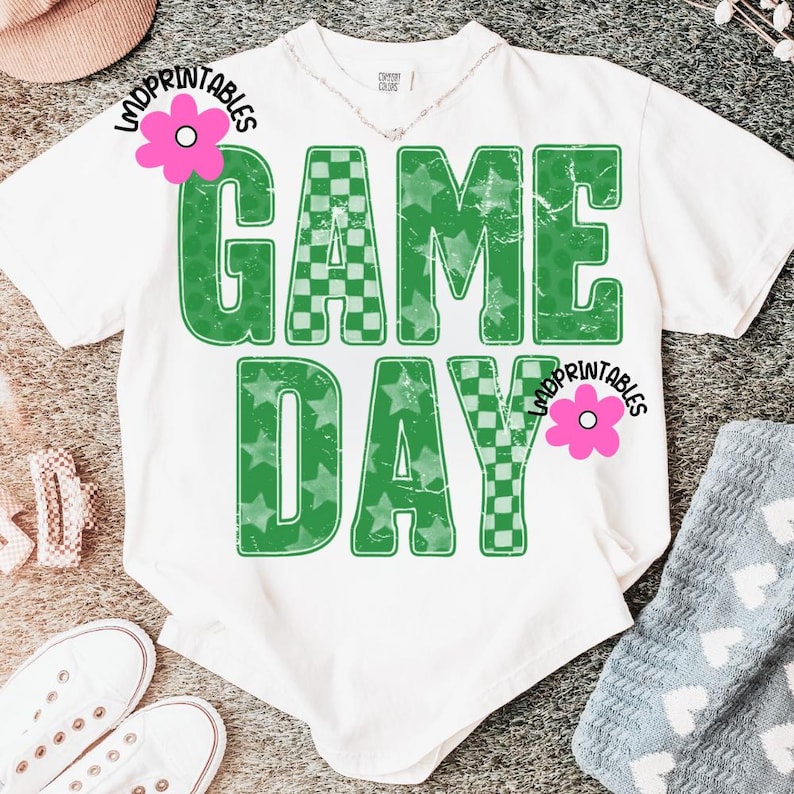 May include: White t-shirt with the words "GAME DAY" in large, distressed green lettering. The letters have a checkerboard and star pattern. The shirt is casual and appears to be made of soft material.