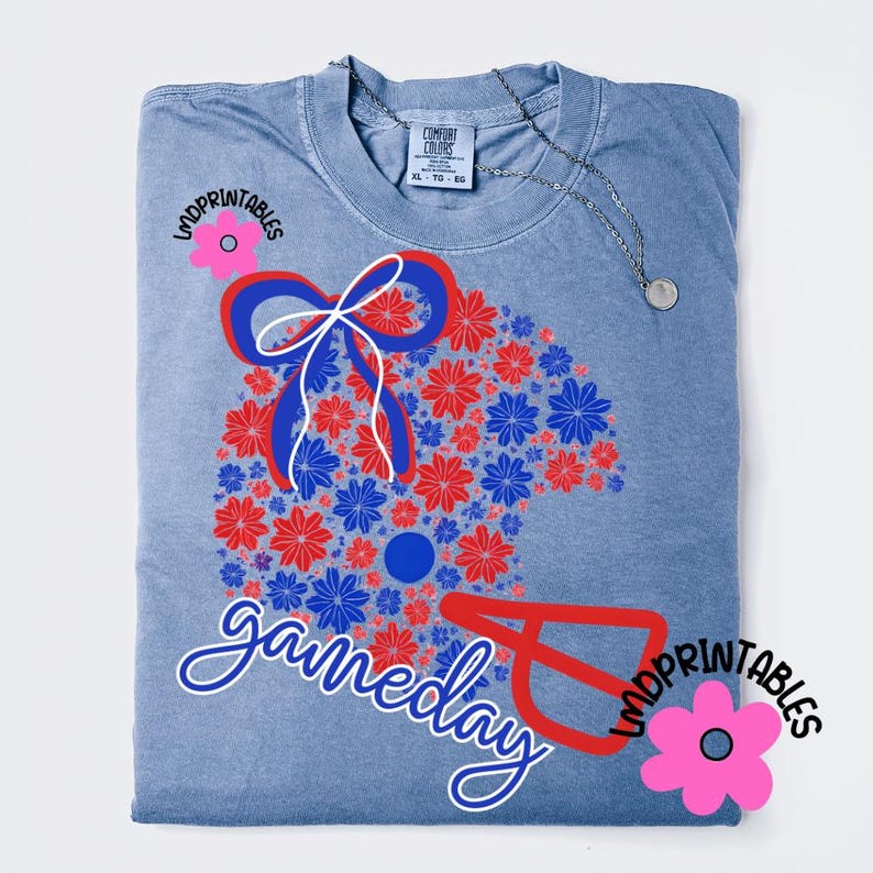 May include: A light blue Comfort Colors t-shirt with a floral design in red and blue, featuring a bow and the word "gameday" in white script. A silver necklace with a pendant is draped on the shirt.